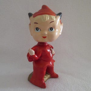 May include: A vintage-style bobblehead figurine featuring a character with a large head, blonde hair, and black pointed ears, wearing a red pointed cap and a matching red outfit. The figure has a pale complexion, painted facial features, and stands.