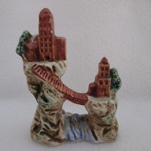 May include: A ceramic figurine depicting a bridge connecting two castle-like structures. The buildings are reddish-brown with detailed windows, set on rocky formations with green and blue accents. A small waterfall is visible.