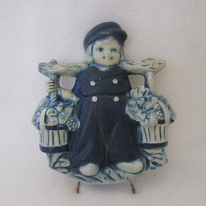 May include: A ceramic wall hanging featuring a Dutch boy carrying baskets of fruit. The figure is dressed in a navy blue outfit with white accents and a matching hat. The baskets and the support beam are light blue.