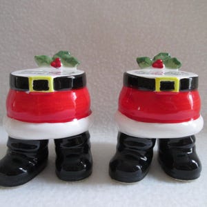 May include: Two ceramic Santa Claus figurines. Each figurine is shaped like Santa's torso and legs. The torso is red with a black belt and gold buckle. The legs are black boots. Each figurine has a white rim and a holly sprig on top.