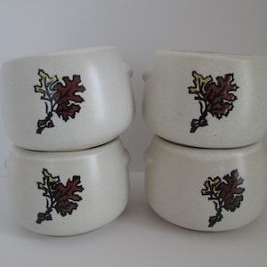 May include: Four off-white ceramic bowls stacked in two pairs. Each bowl features a brown and yellow leaf design. The bowls have small handles on each side. The bowls are likely for soup or cereal.