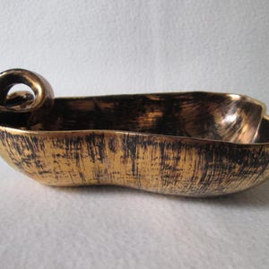 May include: A gold-colored, oblong decorative bowl with a textured surface and a black, distressed finish. The bowl has a looped handle on one end. The interior is a solid gold color.