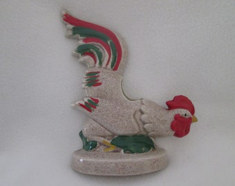 May include: A ceramic rooster planter with a red, green, and white tail. The rooster is sitting on a green leaf and has a red comb.