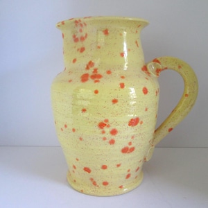 May include: A ceramic pitcher with a glossy pale yellow glaze featuring a speckled pattern of vibrant orange spots. The vessel has a wide, flared rim, a rounded body, and a curved handle attached to the side, creating a functional pouring spout.