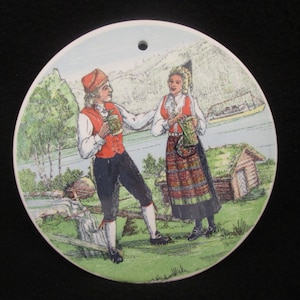 May include: A round ceramic plate with a painted scene of two people in traditional clothing holding mugs, standing in a green field with a river and a house in the background.