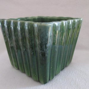May include: Square green ceramic planter with vertical ribbed design. The planter has a glossy finish and a slightly textured surface. The color is a deep, rich green with variations in tone. A decorative piece for indoor or outdoor use.