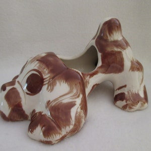 May include: A ceramic planter shaped like a brown and white spotted dog. The dog is lying down with its head turned to the side. The planter has a hole in the top for planting.