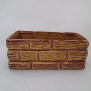 May include: A rectangular ceramic planter with a brick-like design. The planter is painted in shades of brown and tan, with a textured surface that mimics the look of individual bricks. The interior is a darker brown.