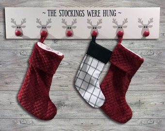 May include: A white wooden stocking holder with six reindeer hooks and the text "~ THE STOCKINGS WERE HUNG ~". Three red stockings are hanging from the hooks, one with a black and white plaid pattern.