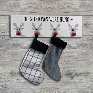 May include: White wooden sign with four reindeer hooks, each with a red nose. The sign reads "~ THE STOCKINGS WERE HUNG ~". Two stockings are hanging from the hooks, one is black and white plaid, the other is gray.