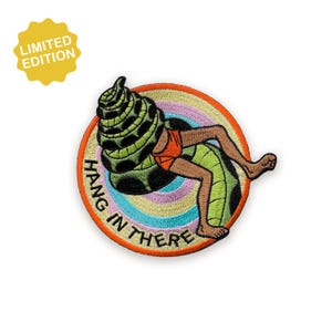 May include: Embroidered patch featuring a cartoon figure emerging from a green snake. The figure wears orange shorts and is surrounded by a rainbow-colored circle. Text reads "HANG IN THERE". A yellow starburst with "LIMITED EDITION" is in the upper left.