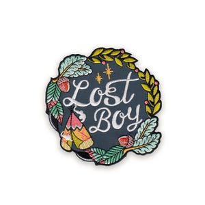 Może przedstawiać: Haftowana naszywka z napisem "Lost Boy" w białym skrypcie na ciemnoniebieskim tle. Naszywka otoczona jest wieńcem z zielonych liści, żółtych jagód, żołędzi i małego tipi.