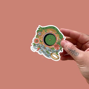 Op de afbeelding: Een sticker met een groene deur, omgeven door baksteen, bloemen en groen. De tekst op de sticker luidt "Not all those who wander are lost" met een gouden ring. De sticker wordt door een hand vastgehouden.