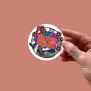 Può includere: Un adesivo rotondo con un'illustrazione di balena, con elementi rossi, blu e floreali, e la scritta "DON'T PANIC". L'adesivo è tenuto in mano su uno sfondo rosa.