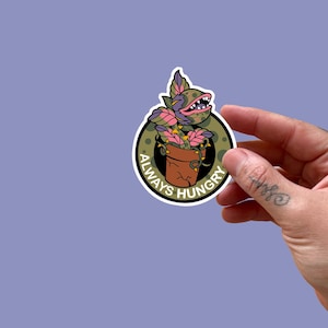 May include: A round sticker featuring a cartoon plant with a toothy grin in a brown pot. The plant has pink and green leaves and is surrounded by a black circle with the words "ALWAYS HUNGRY" in white.