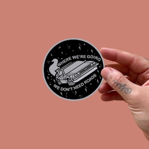 Può includere: Un adesivo rotondo in bianco e nero con un'illustrazione di un'auto DeLorean e la scritta "WHERE WE'RE GOING WE DON'T NEED ROADS". Il design include stelle e una scia di vapore, evocando uno stile retrò-futuristico.