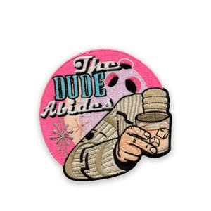 Peut inclure: Écusson brodé avec le texte "The Dude Abides" en turquoise et blanc sur fond rose. L'écusson présente une illustration d'une personne tenant un verre avec des glaçons.