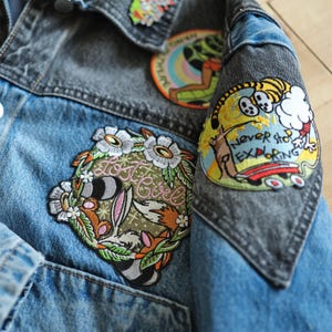 Puede incluir: Primer plano de una chaqueta vaquera con parches. La chaqueta tiene un cuerpo azul claro y mangas grises. Los parches incluyen un dise&ntilde;o floral con un conejo y el texto "Lost Girl", y un tigre de dibujos animados con el texto "Never Stop Exploring".