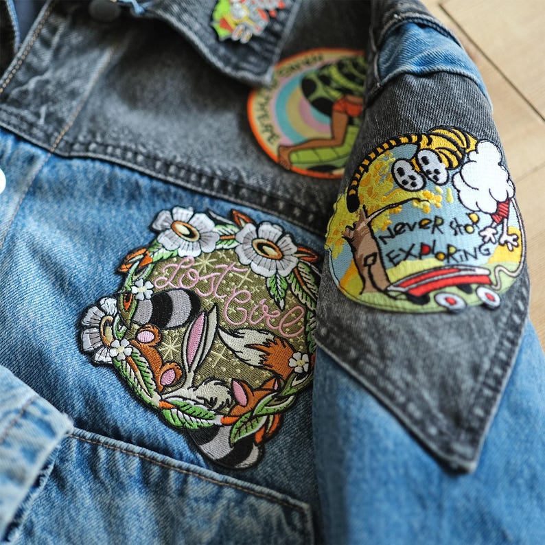 Puede incluir: Primer plano de una chaqueta vaquera con parches. La chaqueta tiene un cuerpo azul claro y mangas grises. Los parches incluyen un dise&ntilde;o floral con un conejo y el texto "Lost Girl", y un tigre de dibujos animados con el texto "Never Stop Exploring".
