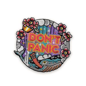May include: Embroidered patch featuring a whale design with the words "DON'T PANIC" in bright yellow and pink. The patch includes pink flowers, clouds, and a colorful ocean scene, all set against a white background.
