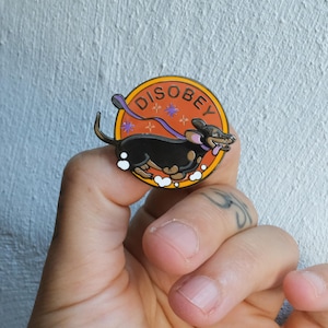 May include: A circular enamel pin featuring a black dachshund dog running against an orange background. The dog has a purple leash trailing behind it, and the word DISOBEY is printed in black letters along the top edge of the orange circle.