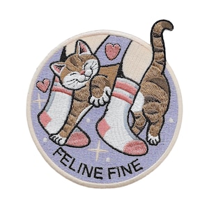 Op de afbeelding: Geborduurde patch met een bruine tabby kat die tegen een witte sok met roze accenten aanleunt. De patch heeft een lichtpaarse achtergrond met de woorden "FELINE FINE" en hart- en kruisdetails.
