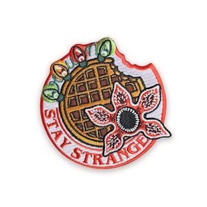 May include: Embroidered patch with a waffle, Demogorgon flower, and string lights. The patch is red, yellow, brown, green, and white, with the words "STAY STRANGE" in white lettering. The design is circular.
