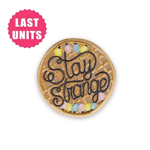 May include: A circular embroidered patch with the words "Stay Strange" in black script on a tan waffle-like background. The patch has a decorative border and colourful lights. A pink circle with "LAST UNITS" is in the upper left.