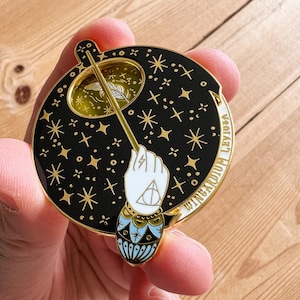 Può includere: Spilla in smalto nero e oro con una bacchetta, una mano e stelle. La mano tiene la bacchetta, con un fulmine e un simbolo triangolare. La spilla ha le parole "Wingardium Leviosa" sul bordo.