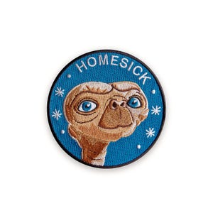 Puede incluir: Parche bordado redondo con el personaje E.T. con ojos azules y cara bronceada. El parche tiene un fondo azul con la palabra "HOMESICK" en blanco. Acentos de estrellas blancas rodean al personaje.