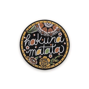 Puede incluir: Parche bordado circular con la frase "hakuna matata" en escritura blanca sobre fondo negro. El parche tiene un borde amarillo y diseños florales en naranja, marrón y amarillo.