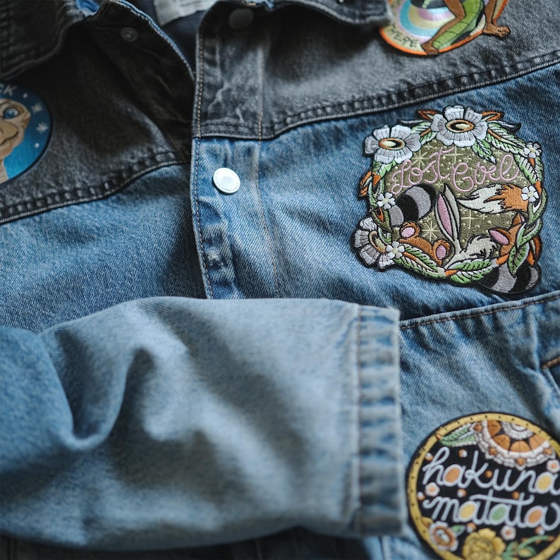 Puede incluir: Chaqueta vaquera azul con varios parches bordados. Los parches incluyen el texto "Lost Girl" rodeado de flores, un personaje de dibujos animados y la frase "Hakuna Matata". La chaqueta es azul y gris.