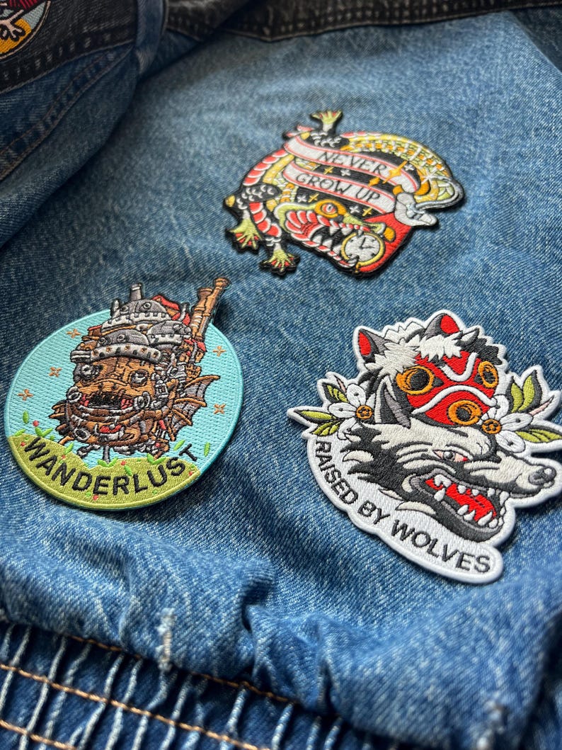 Puede incluir: Tres parches bordados sobre una chaqueta vaquera. Un parche presenta un drag&oacute;n colorido con el texto "NEVER GROW UP". Otro parche tiene un dise&ntilde;o de lobo con el texto "RAISED BY WOLVES". El tercer parche tiene un castillo de dibujos animados con el texto "WANDERLUST".