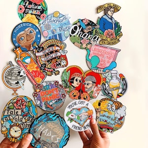 Puede incluir: Una colecci&oacute;n de parches bordados coloridos con varios dise&ntilde;os y texto. Los parches presentan frases como "Hakuna Matata" y "Ohana". Otros parches representan ilustraciones de un dinosaurio, un globo terr&aacute;queo y una pareja.