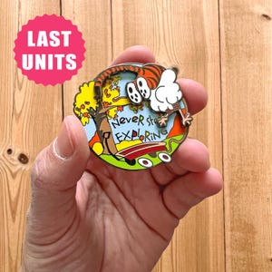 Può includere: Spilla rotonda in smalto con un personaggio a fumetti a forma di tigre e la scritta "Never Stop Exploring". La spilla ha un design colorato con un albero, uno skateboard e uno sfondo di cielo blu. Un adesivo rosa "LAST UNITS" è in alto a sinistra.