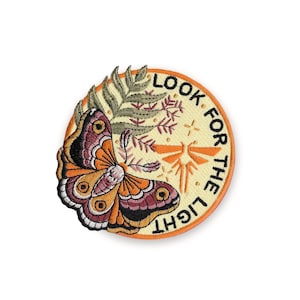 Puede incluir: Parche bordado con una mariposa en tonos naranja, morado y negro, sobre un fondo crema. El parche incluye el texto "LOOK FOR THE LIGHT" en letras negras, con detalles en naranja y un borde decorativo.