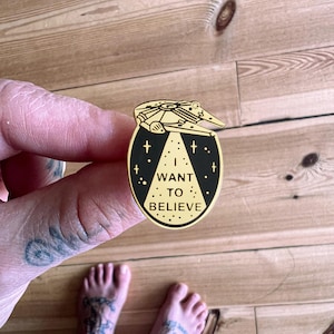 Può includere: Spilla smaltata color oro con un design di astronave sopra un ovale. L'ovale presenta la scritta bianca "I WANT TO BELIEVE" su sfondo nero con stelle e punti bianchi.