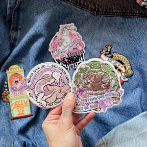 Op de afbeelding: Een hand toont verschillende geborduurde patches met diverse ontwerpen. De patches tonen een eenhoorn, een konijn en een wezen, met tekst als "Fight Against the Sadness" en "Dream Big". De patches zijn op een spijkerjas.