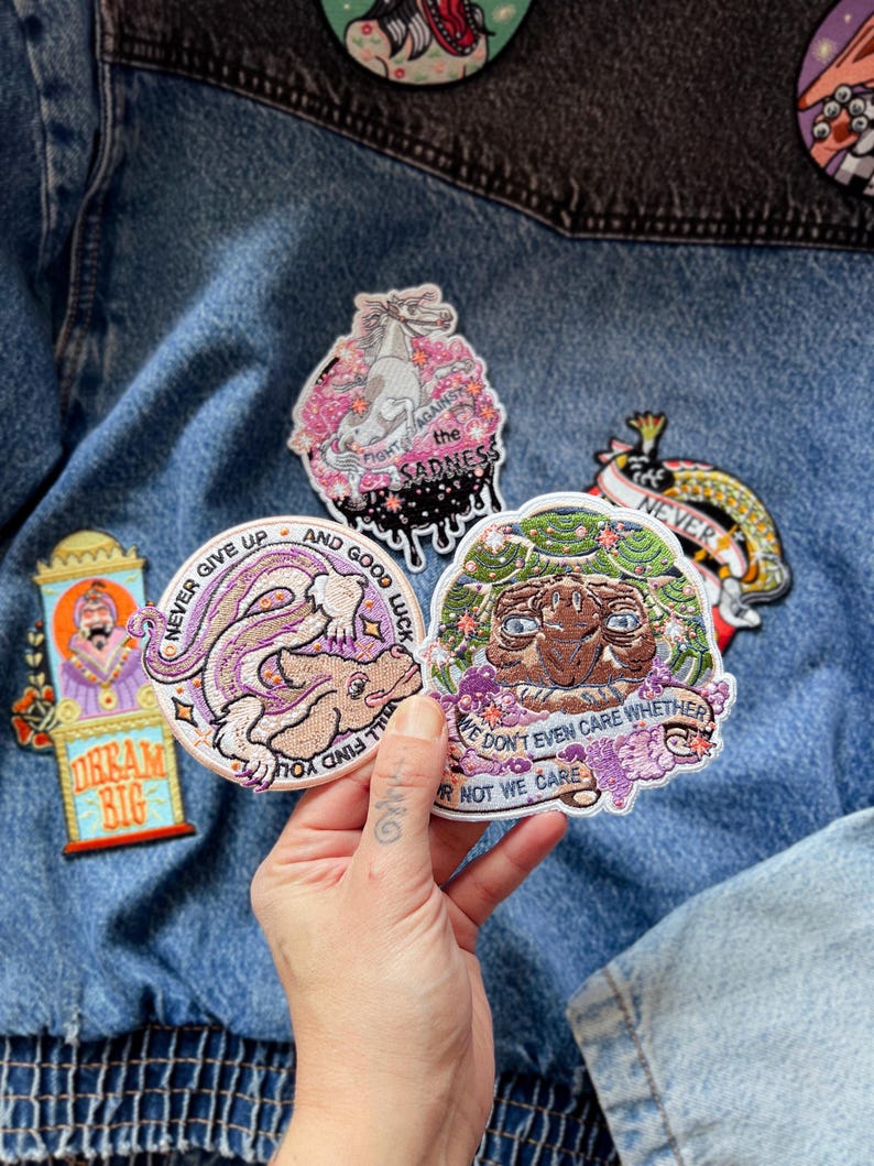 Op de afbeelding: Een hand toont verschillende geborduurde patches met diverse ontwerpen. De patches tonen een eenhoorn, een konijn en een wezen, met tekst als "Fight Against the Sadness" en "Dream Big". De patches zijn op een spijkerjas.