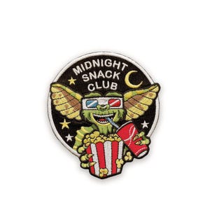 May include: Embroidered patch with a green creature wearing 3D glasses, holding popcorn and a drink. The patch is circular and features the words "MIDNIGHT SNACK CLUB", a crescent moon and stars. The creature has wings and is set against a black background.