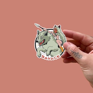 May include: A circular sticker with a cartoon illustration of a person riding a wolf, holding a spear. The person wears a mask and fur clothing. The wolf is gray with a snarling expression. The word "SAVAGE" is written in red.