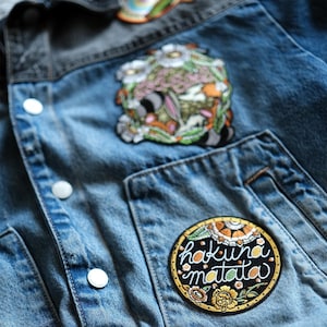 Op de afbeelding: Close-up van een spijkerjasje met drie geborduurde patches. De grootste patch is rond met de woorden "hakuna matata" in witte letters. Andere patches hebben bloemen en abstracte ontwerpen. De jas heeft witte knopen.