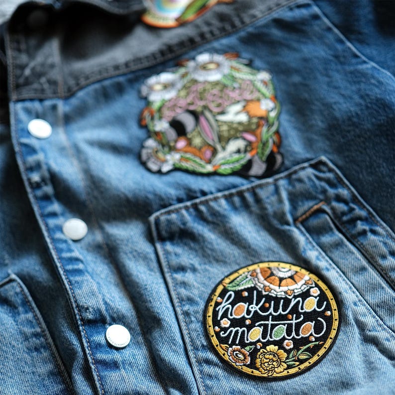 Op de afbeelding: Close-up van een spijkerjasje met drie geborduurde patches. De grootste patch is rond met de woorden "hakuna matata" in witte letters. Andere patches hebben bloemen en abstracte ontwerpen. De jas heeft witte knopen.