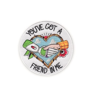 Peut inclure: Écusson brodé avec le texte "YOU'VE GOT A FRIEND IN ME" autour d'un motif en forme de cœur. Le motif représente une poignée de main entre une main et un objet vert et blanc, avec une fleur rouge et un fond de ciel bleu.