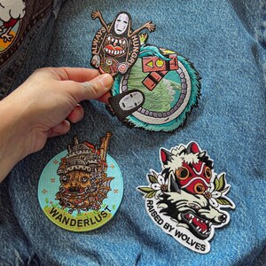 Puede incluir: Parches bordados con dise&ntilde;os inspirados en el anime. Incluye un parche de No-Face con "ALWAYS HUNGRY", una criatura parecida a un drag&oacute;n, un castillo con "WANDERLUST" y un lobo con el texto "RAISED BY WOLVES". Parches sobre una chaqueta vaquera.