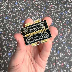 Può includere: Una spilla rettangolare nera e oro con disegni intricati. La spilla presenta un design a cerniera con illustrazioni dettagliate di figure, piante e motivi geometrici. I bordi e le cerniere sono dorati e lo sfondo è nero.