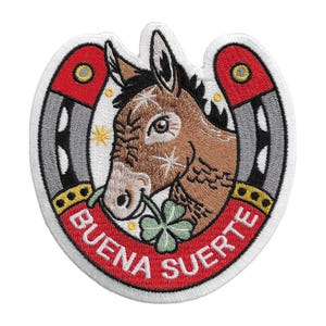 May include: An embroidered patch featuring a brown donkey head centered within a grey and red horseshoe. The donkey holds a green four-leaf clover in its mouth. The red section of the horseshoe contains the white text BUENA SUERTE.