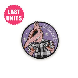 Può includere: Toppa ricamata con un personaggio dei cartoni animati con un lungo naso e una camicia a scacchi. Il personaggio è ambientato su uno sfondo viola con la parola "unusual" in scrittura bianca. Un cerchio rosa con "LAST UNITS" è in alto a sinistra.