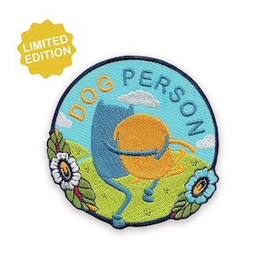 May include: Embroidered patch with a blue and yellow cartoon characters hugging, set against a green landscape with flowers and clouds. The patch reads "DOG PERSON" in yellow text and has a "LIMITED EDITION" tag.