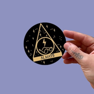 May include: A black circular sticker featuring a gold triangle with a lightning bolt, glasses, and the word "ALWAYS." The design is surrounded by stars. A hand holds the sticker against a light purple background.